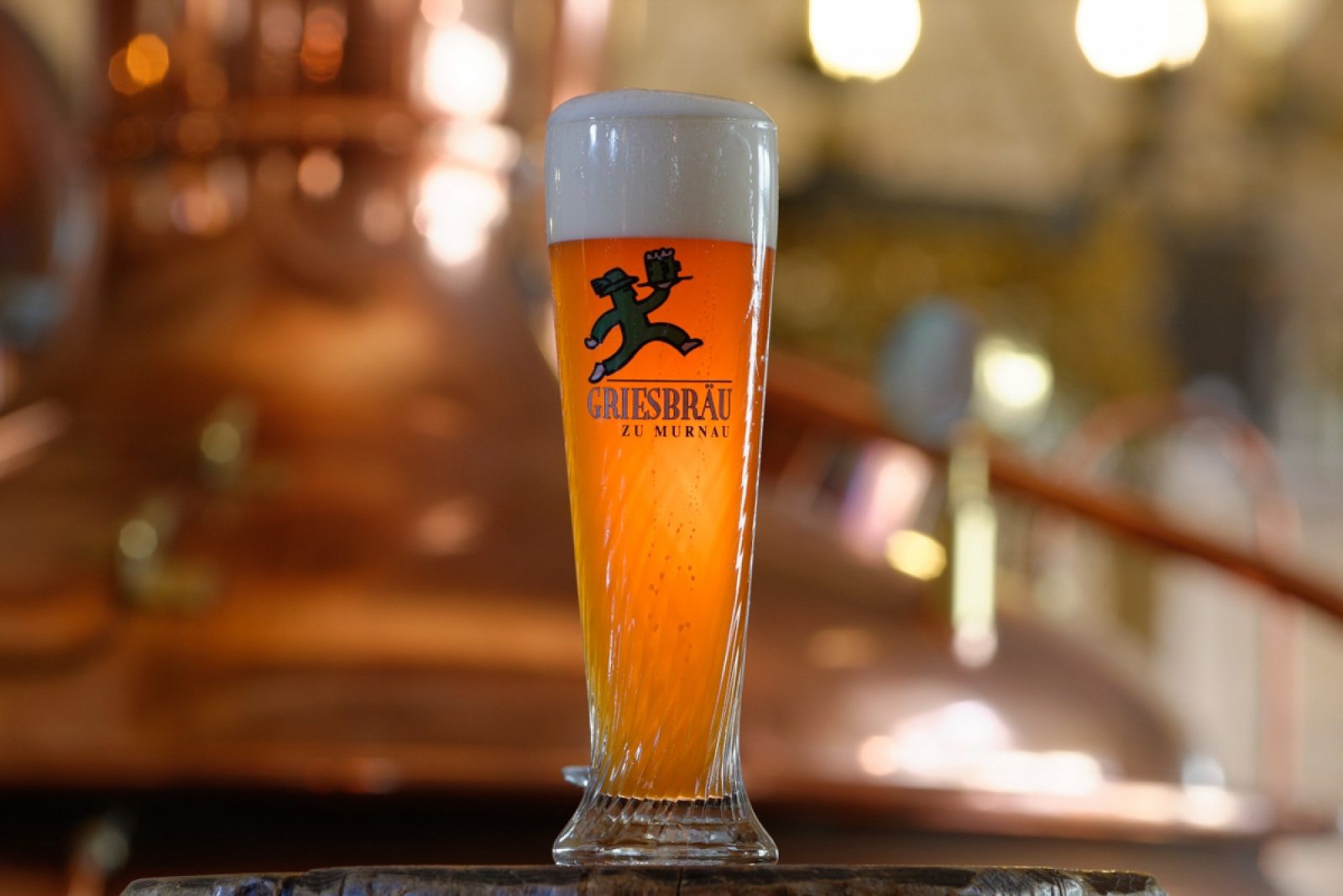 Frisch gezapftes, naturtrübes Griesbräu Weißbier im Glas mit Manschgerl-Logo vor einem kupfernen Braukessel