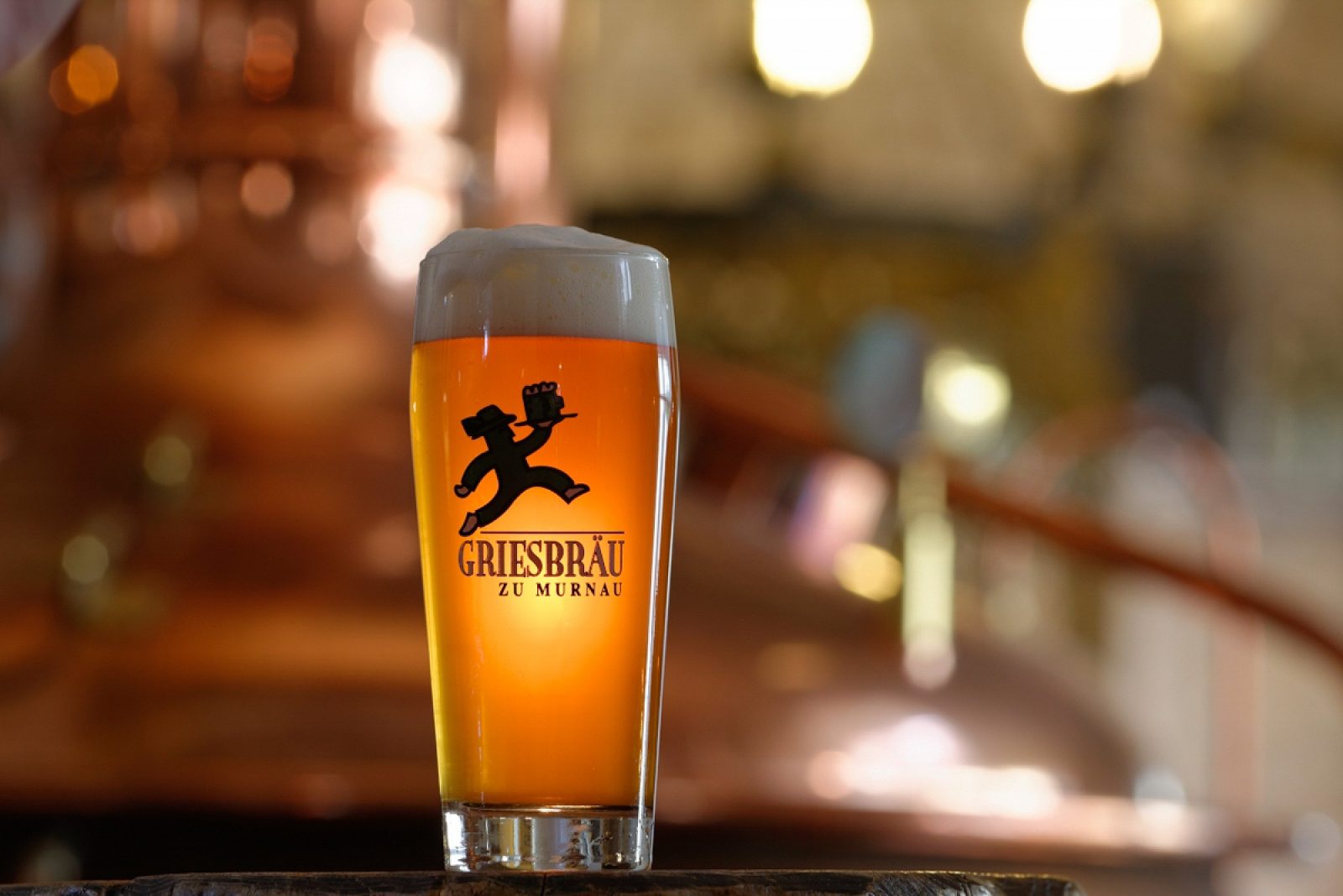 Frisch gezapftes Griesbräu Helles im Glas mit Manschgerl-Logo vor kupfernem Braukessel